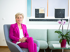 Volksanwältin Gaby Schwarz trägt einen pinken Hosenanzug und sitzt auf einem Polstersessel in ihrem Büro