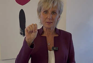 Volksanwältin Gaby Schwarz steht in ihrem Büro und zeigt das SOS Handzeichen bei Gewalt.