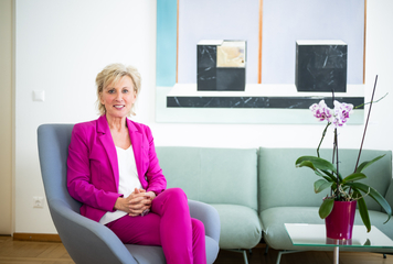 Volksanwältin Gaby Schwarz trägt einen pinken Hosenanzug und sitzt auf einem Polstersessel in ihrem Büro