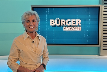 Volksanwältin Gaby Schwarz steht in einem beigen Kleid im ORF Studio vor dem Bürgeranwalt Logo.
